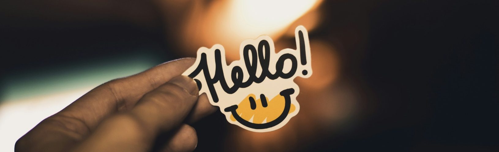 A hand holds a yellow sticker with the text "Hello!" and a smiley face.