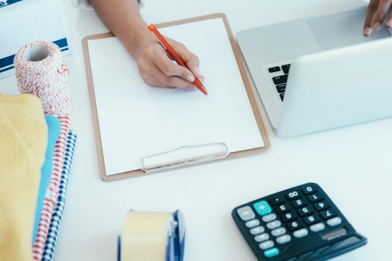 A hand holding a red pen over a blank notepad, with a laptop and calculator nearby.